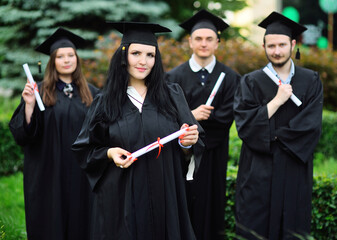 young girl, a university graduate student in a black robe and a square hat with a diploma in her hands, smiles against the background of students ' classmates.
