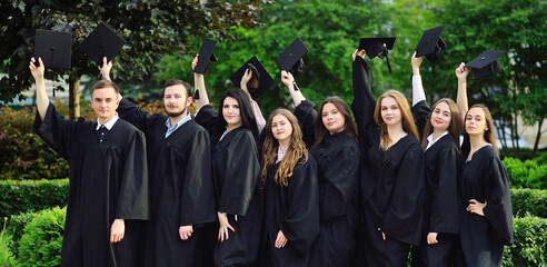 group of university or college graduates in black robes raised their hands with square caps of...