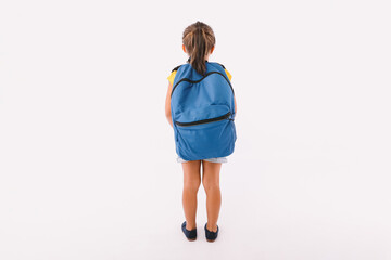 Little girl with black hair dressed in denim overalls and a blue t-shirt, with a backpack ready for going back to school, with her back turned, on white background