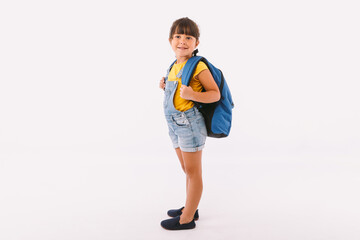 Little girl with black hair dressed in a denim overalls and a blue t-shirt, with a backpack ready for going back to school, on her side, on white background