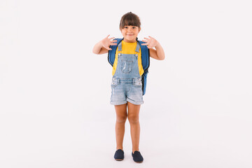 Little girl with black hair dressed in a blue dungarees and a T-shirt, with a backpack ready for back to school, waving with her hands, on white background.