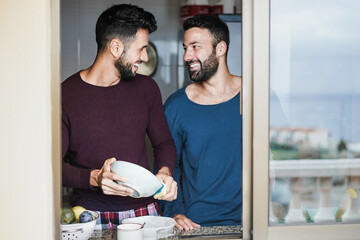 Gay male couple having tender moment while washing dishes inside home kitchen - Focus on left man...