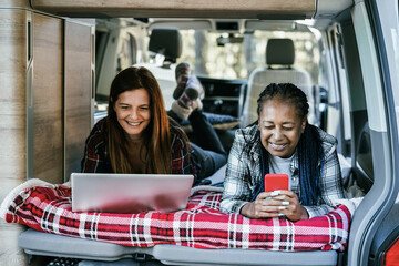 Multiracial women friends having fun camping inside camper van while working with computer laptop...