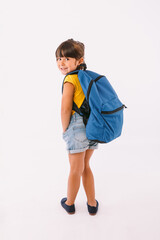 Little girl with black hair dressed in a denim overalls and a blue t-shirt, with a backpack ready for going back to school, on her side, on white background