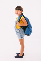 Little girl with black hair dressed in a denim overalls and a blue t-shirt, with a backpack ready for going back to school, on her side, on white background