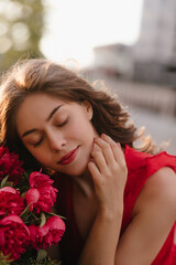 Russian lady in the red beautiful dress touching her soft cheek by hand romantically. Woman in a dreaming mood holding the peony bouquet outside near the cafe. Perfect skin and red lips 