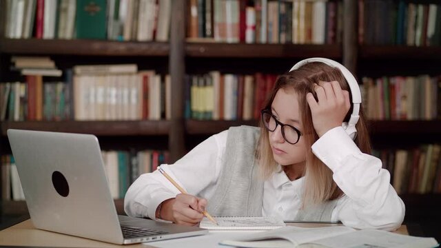 Focused young woman school wear headphones with glasses learn watching online webinar webcast class looking at laptop elearning making notes or video calling remote teacher.