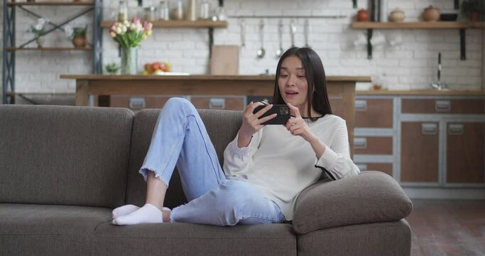 Asian young girl lying in sofa having video chat on smartphone casual conversation at home. Atractive young woman calling, talking on cellphone camera, smiling and laughing. Self isolation concept.