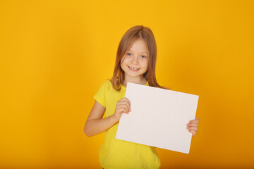 Obraz na płótnie Canvas a beautiful brown-haired girl in a yellow T-shirt holds a white sheet of paper with a yellow background