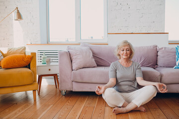 Senior adult smiling woman practicing yoga at home living room. Elderly relaxed female sitting in lotus pose and meditating zen like near sofa