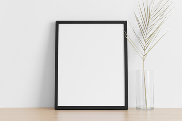 Black frame mockup with a palm leaf on the wooden table.