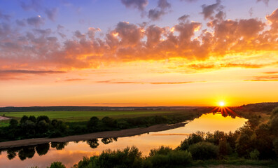 Scenic view at beautiful summer river sunset with reflection on water with green bushes, calm water ,deep colorful cloudy sky and glow on horizon on a background, spring evening landscape