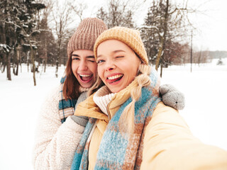 Two young beautiful smiling hipster female in trendy warm clothes and scarfs.Carefree women posing in the street in park. Positive pure models having fun in snow. Enjoying winter moments.Taking selfie