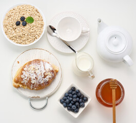 Morning breakfast, raw oatmeal flakes in a ceramic plate, milk in a decanter, blueberries and honey in a jar on a white table