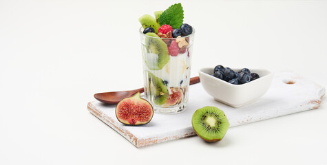 a transparent glass with granola poured with yogurt, on top of ripe raspberries, blueberries and figs on a white table. Healthy breakfast