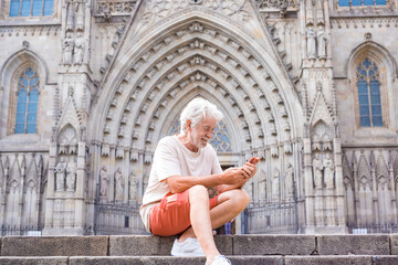 Fototapeta na wymiar Smiling senior white-haired traveler enjoying visit to cathedral of Barcelona, sitting on staircase holding mobile phone. Retired man enjoying free time and journey