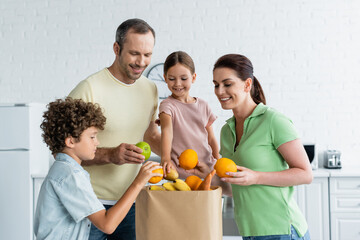 Cheerful family with kids holding fruits near paper bag in kitchen
