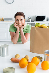 Smiling woman looking at camera near blurred food and paper bag in kitchen