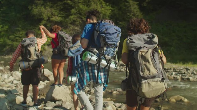 Rear view of positive diverse multiracial tourists with backpacks backpacking by mountain river, overcoming obstacles on the way while enjoying healthy lifestyles and recreation on mountain hiking.
