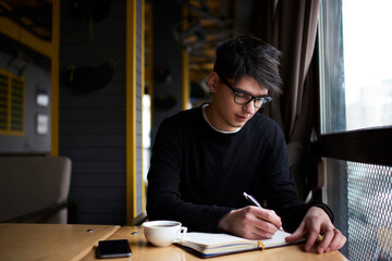 Young smiling millennial male writing notes in notepad while sitting at the table. Financial planning and organizational issues