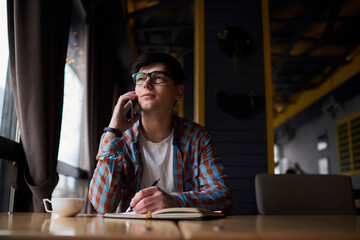 Young freelance worker calling via mobile phone while holding documents in hand. Small owner working remote in coffee shop interior