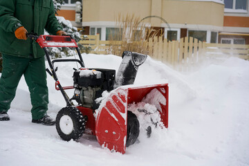 A worker in green overalls removes snow with a red snowblower against the background of a spruce, a snowdrift and a house.