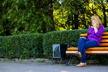 Defocus caucasian blond woman talking, speaking on the phone outside, outdoor. 40s years old woman in purple blouse in park on bench. Adult female using cellphone. Copy space. Out of focus