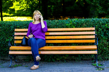 Defocus caucasian blond woman talking, speaking on the phone outside, outdoor. 40s years old woman in purple blouse in park on bench. Adult women using telephone. Copy space. Out of focus