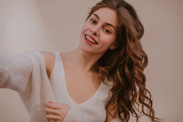 Close-up of fair-skinned lady makes selfie smiling with teeth on light background. Long blond hair scatters on one side. A girl in white T-shirt and warm sweater on top of her shoulders.