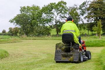A man wearing a high-visibility jacket and ear defenders mows the grass in a large garden while sitting on a ride-on lawn mower