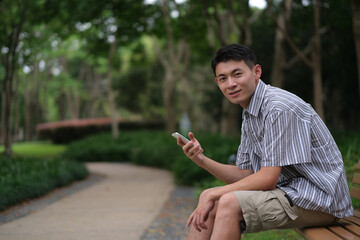 one Asian young man smiling and using phone, looking at camera in natural green park