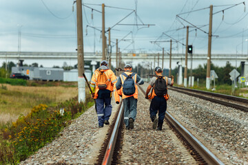 workers in orange uniforms walk along the railway tracks. the repair team is going to work by rail. stone embankment of the railway and power lines. it is dangerous to walk on railway tracks