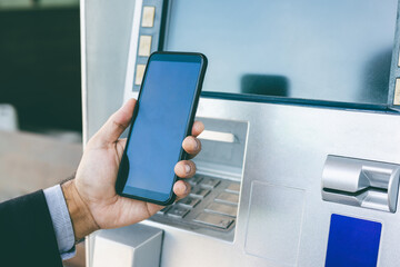 Young businessman standing front of atm machine using smartphone withdraw the money without atm credit card. He ut the pin code into smartphone and take the money.