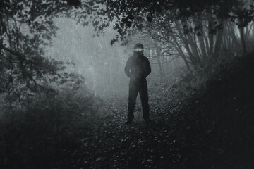 A scary hooded figure with glowing eyes on a path through a moody misty autumn woodland. With a blurred, textured, grunge edit