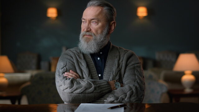 Portrait of senior man sitting at table with crossed hands in vintage cabinet. - Powered by Adobe