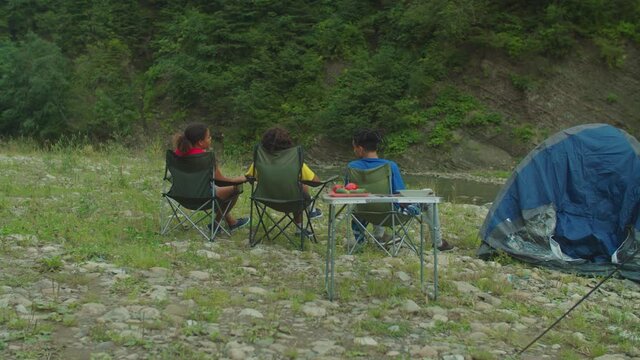 Rear view of happy diverse multiracial tourists sitting on folding chairs, talking and enjoying scenic mountain nature, freedom and leisure while travelers relaxing at camping at mountain riverbank.