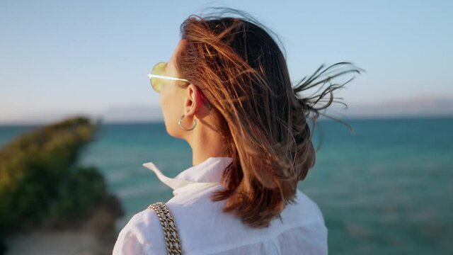 Young woman standing near cliff over Mediterranean Ionian sea. Her hair flutter beautifully. Lady watching beautiful blue water surface alone, nature background. Windy weather, golden hour.