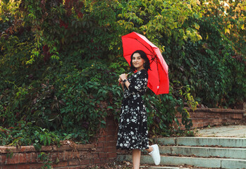 A girl in a black dress stands in the park under a red umbrella