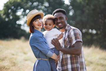 Portrait of cheerful multicultural parents hugging little son from both sides while posing on...