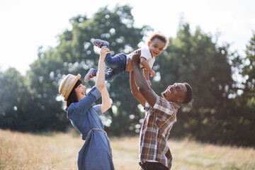 Side view of young parents smiling sincerely while playing with their little son outdoors. Caucasian mother and african father spending time with lovely child on summer nature.