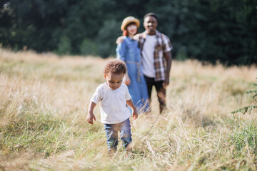 Fototapeta na wymiar Pretty african boy running and playing among high grass while his multiracial parents standing in hugs on blur background. Concept of family, happiness and lifestyles.