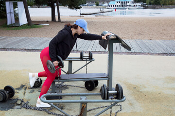 Young beautiful sportive girl performs exercises with dumbbells. Fitness on the street.