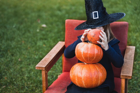 Scary little girl in witch costume, hat with many pumpkins celebrating halloween holiday. Sitting on armchair in coat with pumpkin. Stylish image. Horror, fun at children's party in barn on street