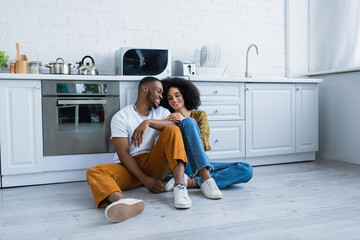 African american man sitting near smiling girlfriend on floor in kitchen.