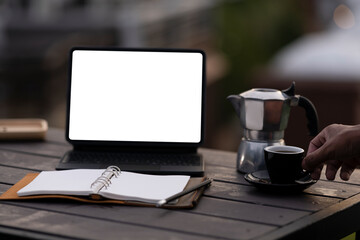 Photo of a white blank screen computer tablet putting on a wooden table surrounded by steel kettle, notebook, and coffee cup over the natural outdoors as a background.