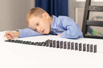 Six year old cute boy plays dominoes at home on a white wooden table. Selective focus. Close-up
