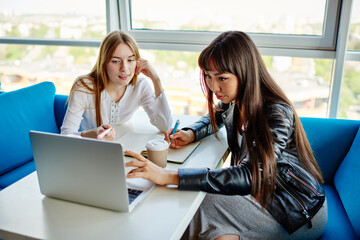 Asian and Caucasian female freelancers collaborating on web project using laptop for watching video webinar, skilled diverse women browsing web page and searching education information for studying