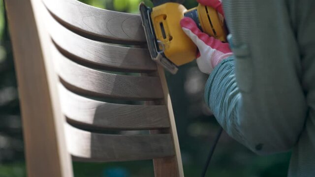 Closeup detail of woman holding electric sander sanding a teak chair outside with dust.