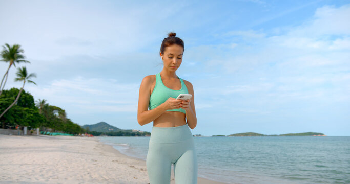 Cute brunette woman walking on sandy beach after her fitness exercise, she types massage and smiles
