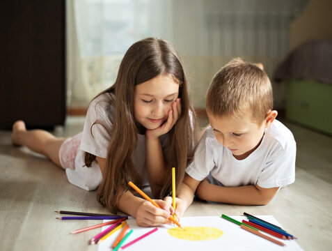 a girl and a boy lie on the floor in the room and draw a picture on a white sheet of paper with colored pencils